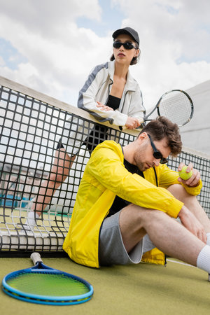 sporty couple in sunglasses posing near tennis net, holding rackets and ball, active wear fashionの写真素材