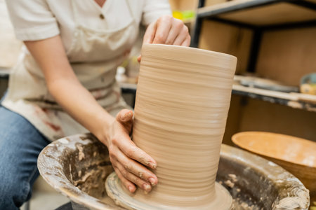 Cropped view of blurred craftswoman in apron creating clay vase on pottery wheel in workshopの写真素材