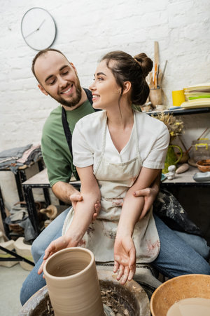 Positive artisan hugging girlfriend in apron and making clay vase on pottery wheel in studioの写真素材