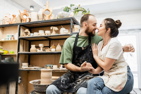 Side view of couple of artisans in aprons holding hands and smiling near pottery wheel in studioの写真素材