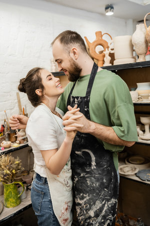 Smiling couple of artisans in aprons holding hands and hugging in ceramic workshopの写真素材