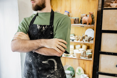 Cropped view of bearded artisan in apron crossing arms while standing in ceramic workshop