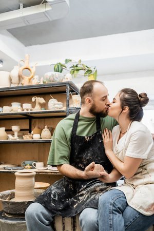 Couple of sculptors in aprons holding hands and kissing near pottery wheel in workshopの写真素材