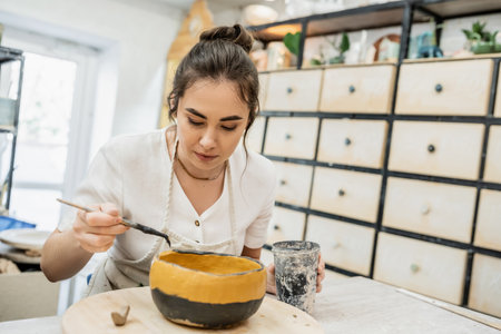 Brunette craftswoman in apron coloring ceramic bowl while working in pottery studio at background