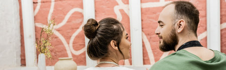 Bearded craftsman looking at brunette girlfriend near clay in pottery workshop, banner