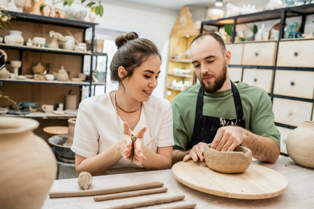 Smiling craftswoman in apron molding clay while boyfriend making bowl in ceramic workshopの写真素材