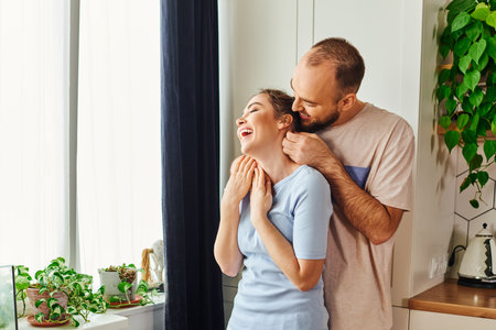 Positive bearded man in homewear hugging cheerful girlfriend in kitchen at homeの写真素材
