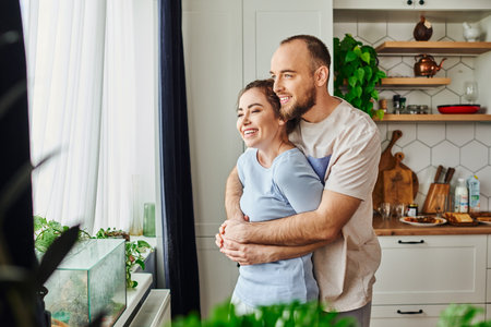 Positive man in homewear hugging brunette girlfriend while standing near plants and window at homeの写真素材