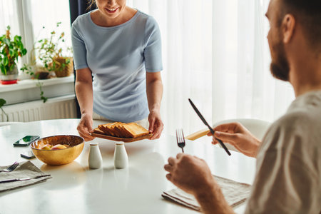 Smiling woman putting toasts on table near boyfriend with cutlery and smartphones in morning at homeの写真素材