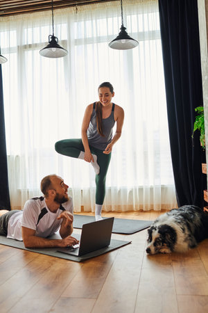 Positive woman standing on fitness mat near boyfriend using laptop and border collie at homeの写真素材