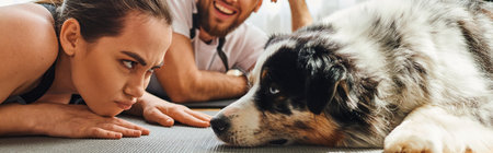 Displeased woman looking at border collie near smiling boyfriend on fitness mat at home, banner