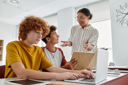 Smiling african american teacher talking to teen schoolboys during lesson with devices in school