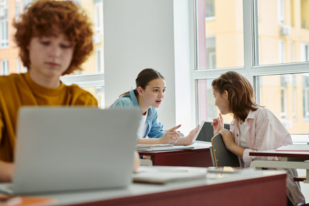 Teenage classmates talking and pointing with fingers near devices and notebook in classroom