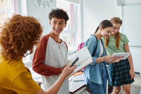 Smiling schoolboy with braces holding notebook near friends with devices in classroom in schoolの写真素材