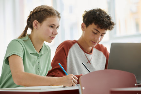 Teenage schoolgirl writing on notebook near blurred classmate and laptop during lesson in classの写真素材