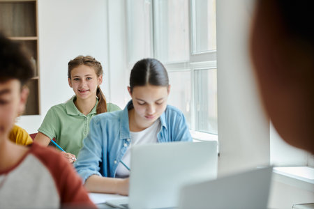 Teen schoolgirl looking at camera and smiling near blurred classmates during lesson in classroom