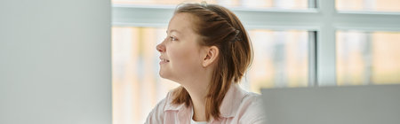 Side view of smiling teen schoolgirl looking away near blurred laptop during lesson in class, banner