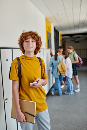 redhead schoolboy holding smartphone, happy student in school hallway looking at camera during break