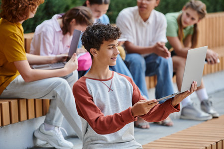 teenage boy using laptop near classmates and teacher, blur, diversity, smile, e-study, educationの写真素材