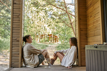 Side view of positive romantic couple sitting near wine on porch of wooden summer house outdoorsの写真素材