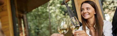 Happy brunette woman toasting wine with boyfriend while sitting in hammock near summer house, bannerの写真素材
