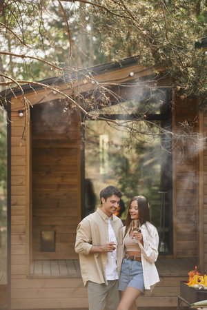 Cheerful and stylish couple holding wine and standing near grill with smoke and vacation houseの写真素材