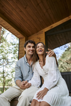 man pointing away and hugging cheerful woman while sitting on porch, happy couple, vacation houseの写真素材