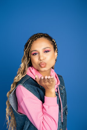 beautiful african american woman sending air kiss on blue background, denim vest and pink hoodieの写真素材