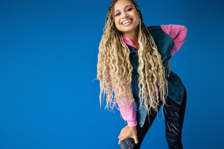 happy african american woman in pink hoodie and denim vest posing on blue background, dreadlocksの写真素材