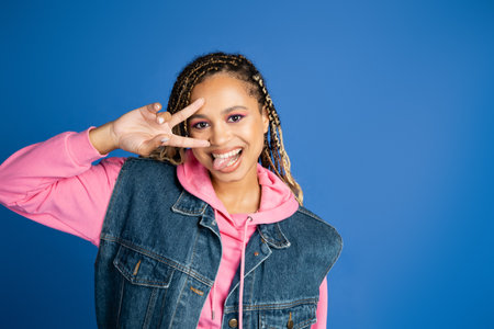 happy african american woman with dreadlocks showing v sign with hand, sticking out tongue on blueの写真素材