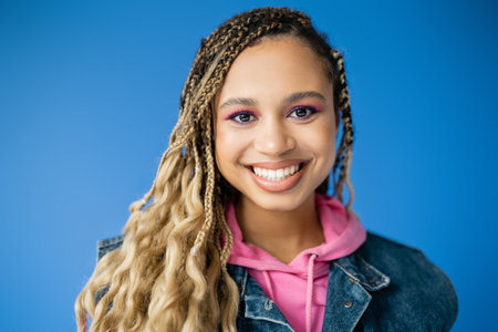joyful african american woman in pink hoodie and denim vest on blue background, dark skinned modelの写真素材