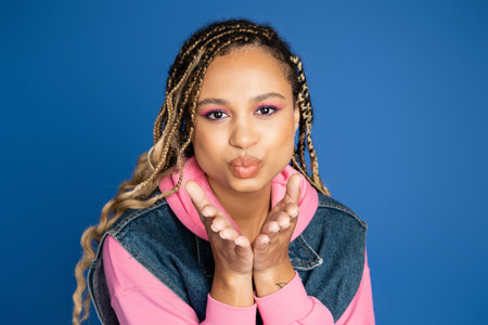 attractive african american woman sending air kiss on blue background, denim vest and pink hoodieの写真素材