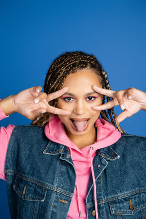 african american woman with dreadlocks showing v sign with hands and sticking out tongue on blue