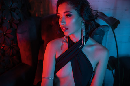 Portrait of sexy young asian woman in dress looking away near plants in neon light in night clubの写真素材