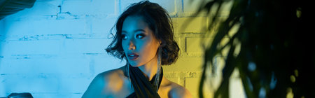 Stylish young asian woman looking away and standing near plants in neon light in night club, banner