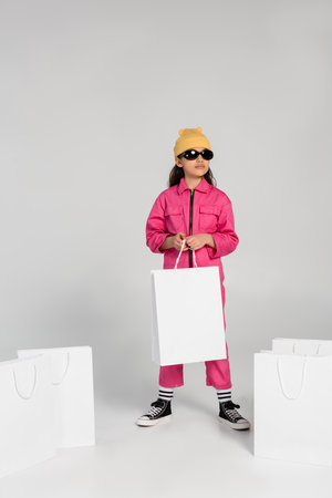 stylish preteen girl in beanie hat and sunglasses standing and holding shopping bags on greyの写真素材