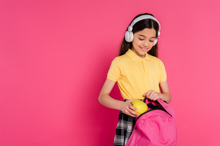 happy schoolgirl in wireless headphones putting apple inside of backpack, pink background, studentの写真素材