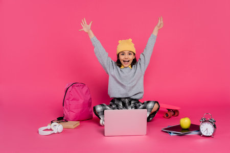 cheerful girl in beanie hat using laptop, sitting near headphones, apple, backpack, alarm clockの写真素材