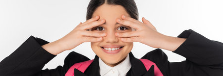 happy schoolgirl covering eyes isolated on white, student in uniform, looking at camera, bannerの写真素材