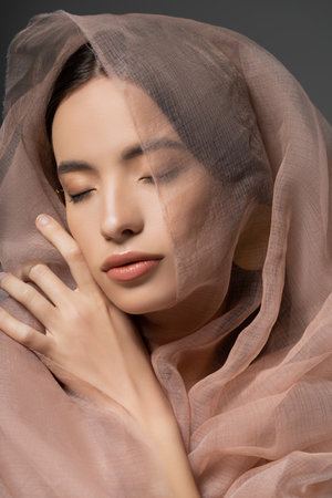 Portrait of beautiful young asian woman with natural makeup posing with beige cloth isolated on greyの写真素材