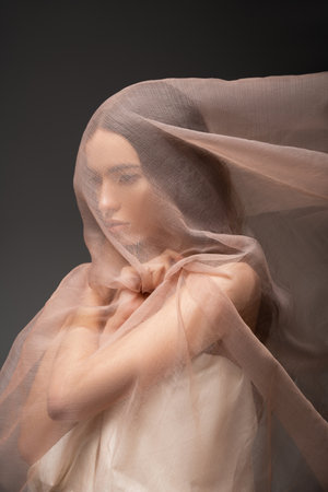 Brunette asian woman looking away while posing and touching beige fabric isolated on blackの写真素材