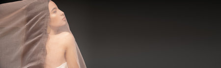Side view of young asian woman with naked shoulder posing under fabric isolated on black, banner