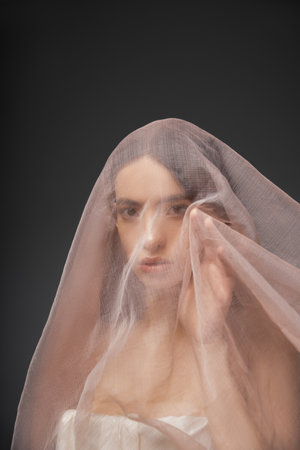 Young asian model looking at camera while posing under beige cloth isolated on blackの写真素材