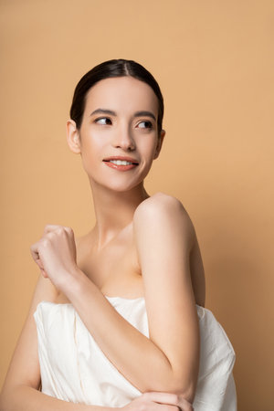 Smiling young asian woman in top with naked shoulders standing isolated on beigeの写真素材