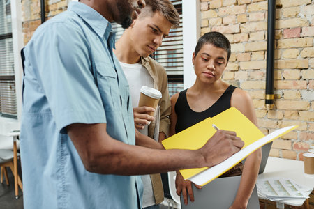 african american man taking notes, writing on paper, holding folder near colleagues in coworkingの写真素材