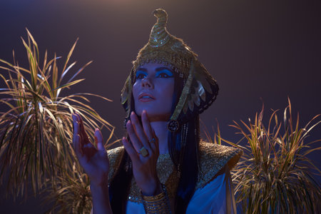 Woman in egyptian costume looking away near desert plants in blue light on brown backgroundの写真素材