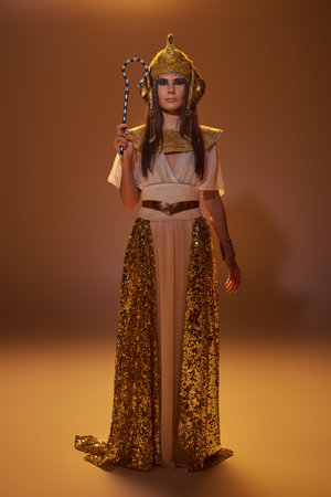 Full length of stylish woman in egyptian attire holding crook and standing on brown backgroundの写真素材