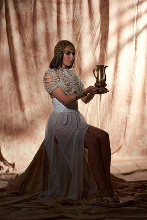 Woman in traditional egyptian outfit holding golden jug while sitting on abstract backgroundの写真素材