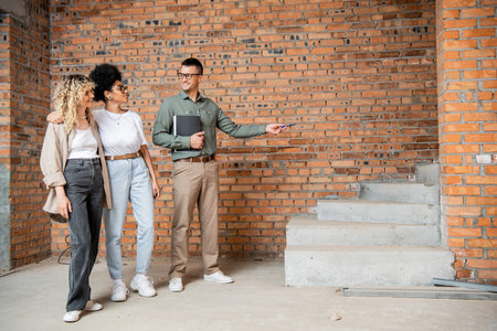 smiling real estate broker pointing at stairs while showing new house to lesbian multiethnic coupleの写真素材