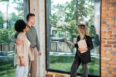real estate agent with folder pointing with hand at window in new cottage near interracial couple